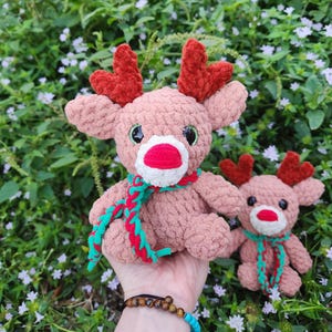 May include: Two crocheted reindeer toys with red antlers, pink bodies, and green and red scarves. The larger reindeer is being held in a hand.