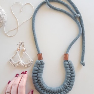 Puede incluir: Collar de cuerda azul grisáceo hecho a mano con detalles de madera marrón. También se ven pendientes a juego de color crema con ganchos plateados. Una bola de hilo blanco y un corazón hecho de hilo están en el fondo.