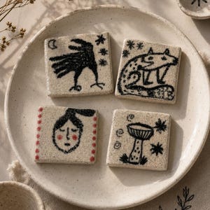 Może przedstawiać: Cztery kwadratowe płytki ceramiczne z nakrapianym beżowym wykończeniem, każda z ręcznie rysowanymi czarnymi ilustracjami przedstawiającymi wronę, cętkowane stworzenie, twarz z czerwonymi akcentami oraz grzyb, ułożone na pasującym okrągłym talerzu.