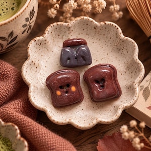 Puede incluir: Tres botones pequeños y brillantes con forma de tostada en un plato de cerámica moteado con bordes festoneados. Dos botones son de color marrón oscuro con detalles naranjas y uno es de color morado oscuro. Cada botón tiene dos agujeros.