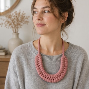 May include: A chunky, braided necklace in a dusty rose color. The necklace features a thick, rounded, woven cord design that sits in a U-shape around the neck, connected to a simple, thin cord that extends upward to form the necklace loop.