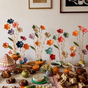 May include: A collection of ceramic flower sculptures on thin wire stems, featuring multi-colored petals in shades of blue, orange, pink, and red. The flowers are arranged in small containers, including halved oranges, alongside various breads and vegetables.