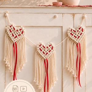 Peut inclure: Une guirlande en macramé composée de trois cœurs blanc cassé tissés avec des rubans rouges et des franges. Les cœurs sont reliés par une corde et incluent un badge circulaire avec le texte PDF & VIDEO TUTORIAL.
