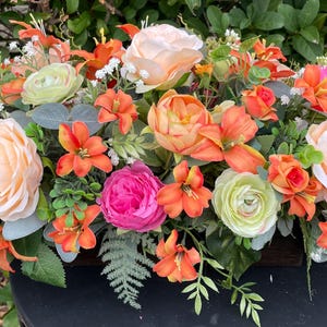 May include: A floral arrangement in a wooden box with a variety of flowers, including peach roses, pink roses, orange lilies, and green ranunculus. The arrangement is set against a backdrop of green foliage.