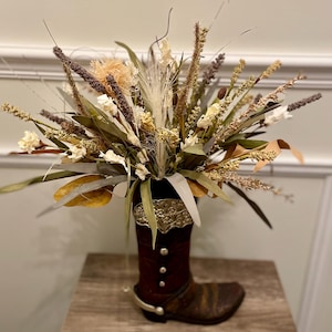 May include: A decorative floral arrangement in a brown cowboy boot vase. The arrangement features dried flowers and grasses in shades of beige, brown, and green. The boot has silver accents and is placed on a wooden surface, creating a rustic, country-themed display.