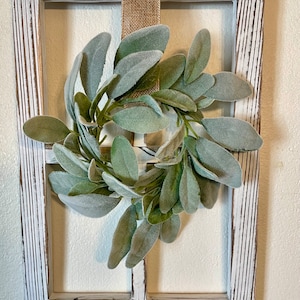 May include: A whitewashed wooden window frame with a rustic, distressed finish. A green faux lamb's ear wreath hangs in the center of the frame, secured with a burlap ribbon.