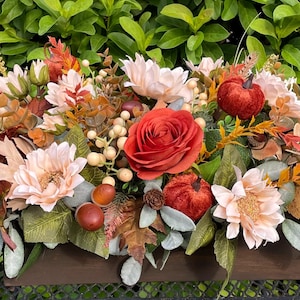 May include: A fall-themed floral arrangement in a wooden trough. The arrangement features red roses, peach-colored dahlias, small red pumpkins, and various fall foliage. The background is a lush green bush.