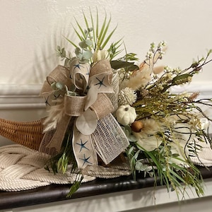 May include: A woven cornucopia centerpiece filled with dried flowers, greenery, and small white pumpkins. A large burlap bow with blue starfish accents adds a decorative touch. The arrangement sits on a cream-colored braided rope mat.