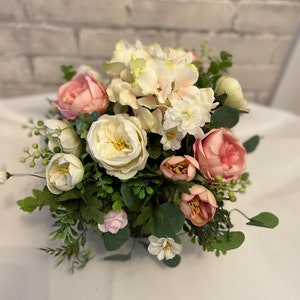 May include: A floral arrangement in a white vase with pink and white flowers, including roses, hydrangeas, and greenery.
