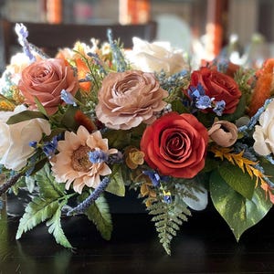 May include: A fall-themed centerpiece with a variety of flowers in shades of red, orange, and white. The centerpiece is arranged in a rectangular container and features greenery, ferns, and other foliage.
