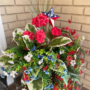 May include: A decorative floral arrangement in a dark pot, featuring red geraniums, blue and white flowers, and greenery. A patriotic star decorates the arrangement. The pot is placed in front of a brick wall.