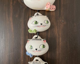 May include: A set of four white ceramic cat-shaped spice jars with green eyes and pink accents. Each jar is labeled with a spice name: garlic salt, paprika, cinnamon, and nutmeg. The jars have colorful bow accents.