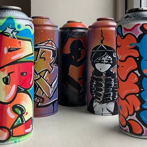 May include: Five spray paint cans with different colorful designs. The cans are all the same size and shape. The designs include a cartoon character, a person with a jacket, and abstract shapes.