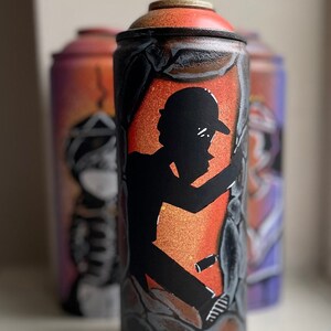 May include: A black and orange spray paint can with a silhouette of a person wearing a hat and holding a spray can. The can is decorated with a black and white abstract design.