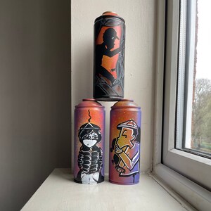 May include: Three spray paint cans with different designs. The top can has a black silhouette of a person with a hat. The middle can has a black and white illustration of a person with a hat and a jacket. The bottom can has a colorful illustration of a person with a hat and a shirt.