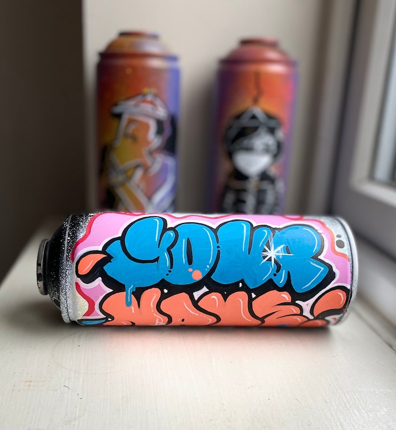 May include: A pink and white spray paint can with blue and orange graffiti art. The word "COVER" is written in large blue letters.