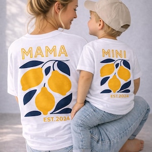 Op de afbeelding: Witte T-shirts met bijpassende citroenafbeeldingen. Op één shirt staat "MAMA" en op de andere "MINI", beide in gouden letters. Het citroenontwerp bevat gele citroenen en marineblauwe bladeren, met "EST.2024" eronder.