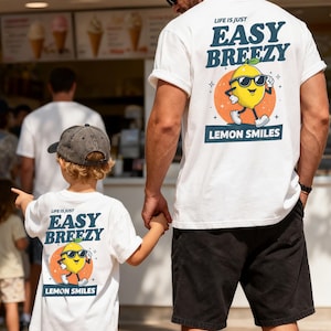 May include: White t-shirts with the text "LIFE IS JUST EASY BREEZY" and a cartoon lemon wearing sunglasses. The lemon is on an orange circle with the words "LEMON SMILES" below. The shirts are worn by a man and a child.