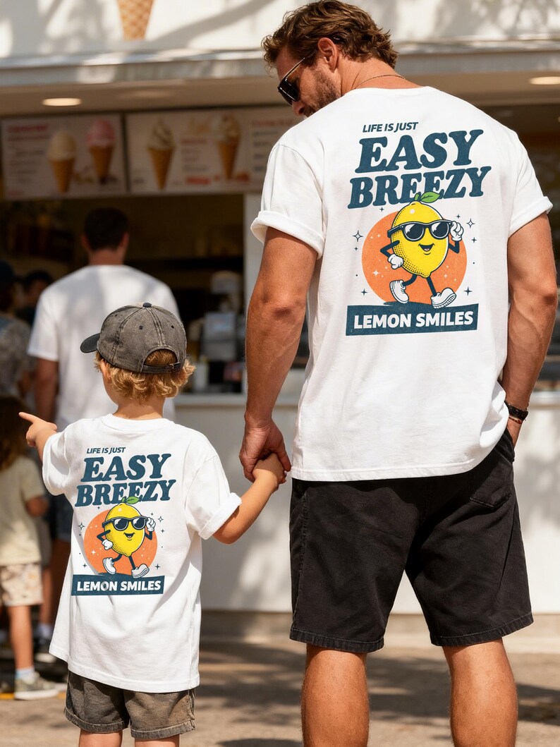 May include: White t-shirts with the text "LIFE IS JUST EASY BREEZY" and a cartoon lemon wearing sunglasses. The lemon is on an orange circle with the words "LEMON SMILES" below. The shirts are worn by a man and a child.