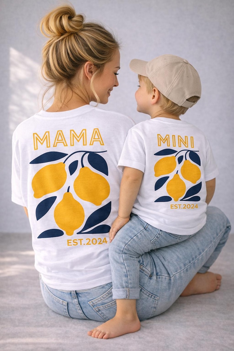 Może przedstawiać: Białe koszulki z pasującymi wzorami graficznymi z cytrynami. Jedna koszulka ma napis "MAMA", a druga "MINI" złotymi literami, z motywem cytryny i liści. Koszulki zawierają r&oacute;wnież napis "EST.2024" w kolorze złotym. Matka i dziecko noszą koszulki.