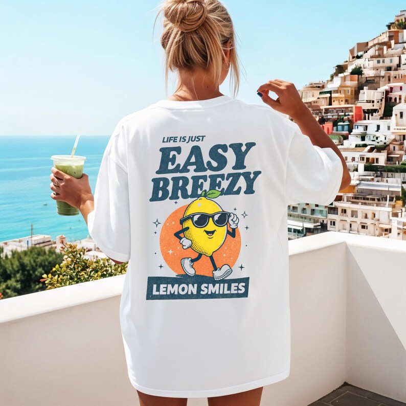 May include: White t-shirt with the text "LIFE IS JUST EASY BREEZY LEMON SMILES" and a cartoon lemon wearing sunglasses. The lemon is set against an orange circle with a blue sky and ocean background.