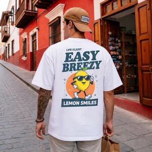May include: White t-shirt with a graphic of a lemon wearing sunglasses and the text "LIFE IS JUST EASY BREEZY LEMON SMILES". The shirt has a relaxed fit and short sleeves. The person is wearing a brown baseball cap.
