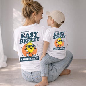 May include: White t-shirts with matching designs. The shirts feature a cartoon lemon wearing sunglasses with the text "LIFE IS JUST EASY BREEZY LEMON SMILES". The shirts are worn by a person and a child.