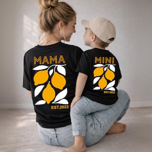 Może przedstawiać: Czarne koszulki z pasującymi wzorami graficznymi z cytrynami. Jedna koszulka ma napis "MAMA", a druga "MINI", obie z napisem "EST.2023" pod wzorem cytryny. Koszulki noszą matka i dziecko.