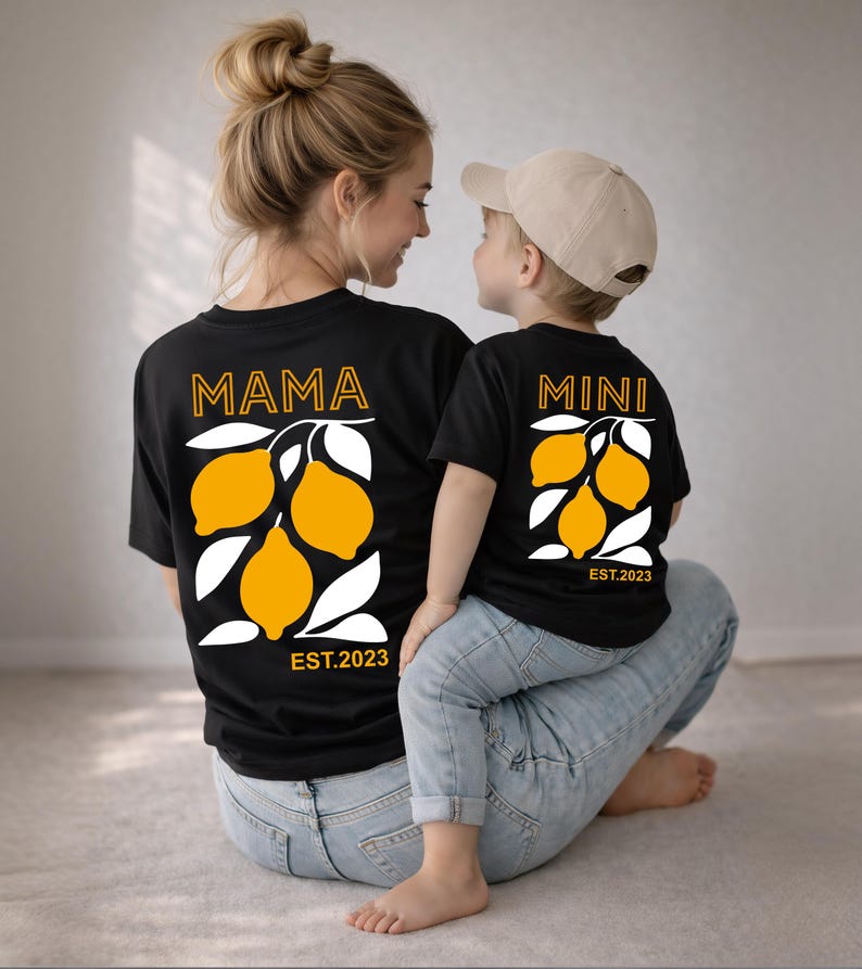 Może przedstawiać: Czarne koszulki z pasującymi wzorami graficznymi z cytrynami. Jedna koszulka ma napis "MAMA", a druga "MINI", obie z napisem "EST.2023" pod wzorem cytryny. Koszulki noszą matka i dziecko.