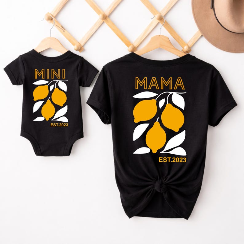 Może przedstawiać: Czarne koszulki z pasującymi wzorami. Jedna koszulka ma napis "MAMA", a druga "MINI". Obie mają ż&oacute;łtą grafikę cytryny z białymi liśćmi i napisem "EST.2023".