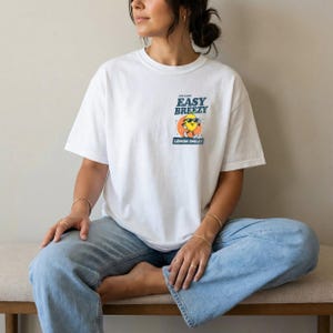 May include: White t-shirt with a graphic of a smiling lemon wearing sunglasses, the text "Easy Breezy Lemon Smiles" is above the lemon. The shirt is a short-sleeved, casual style.