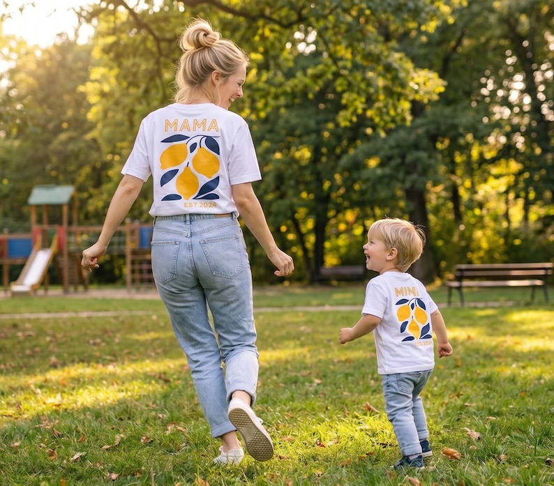 Może przedstawiać: Kobieta i dziecko spacerują po parku. Oboje mają na sobie białe koszulki z motywem cytryny. Na koszulce kobiety napisane jest "MAMA", a na koszulce dziecka "MINI". Mają na sobie niebieskie dżinsy.