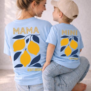 Może przedstawiać: Jasnoniebieskie koszulki z grafiką cytryn. Jedna koszulka ma napis "MAMA", a druga "MINI" w złotym kolorze. Obie mają wz&oacute;r gałązki cytryny i napis "EST. 2024". Komplet pasujących koszulek.