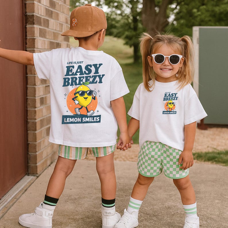 May include: Two children wearing white t-shirts with a lemon graphic and the text "Easy Breezy Lemon Smiles." One child wears striped shorts, the other checkered shorts. Both wear sunglasses and socks.