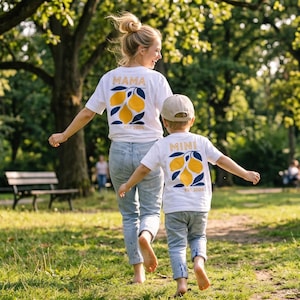 Może przedstawiać: Dwie białe koszulki z pasującymi wzorami cytryn i napisami "MAMA" i "MINI" złotymi literami. Koszulki mają r&oacute;wnież napis "EST 2024". Kobieta i dziecko noszą koszulki podczas spaceru w parku.