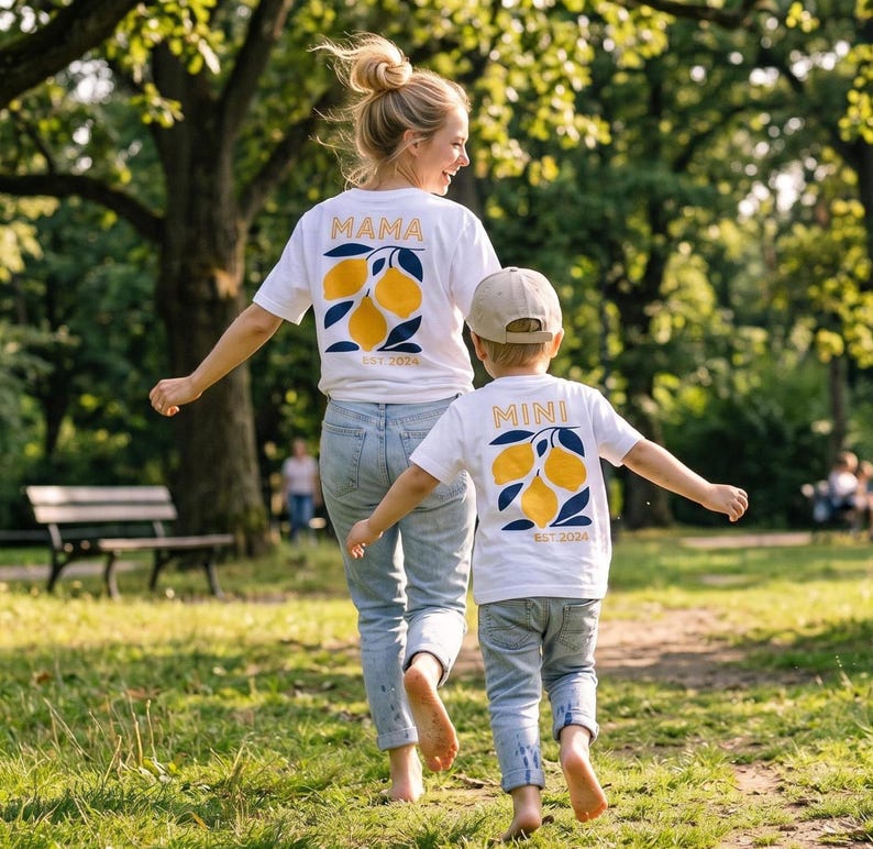 Może przedstawiać: Dwie białe koszulki z pasującymi wzorami cytryn i napisami "MAMA" i "MINI" złotymi literami. Koszulki mają r&oacute;wnież napis "EST 2024". Kobieta i dziecko noszą koszulki podczas spaceru w parku.