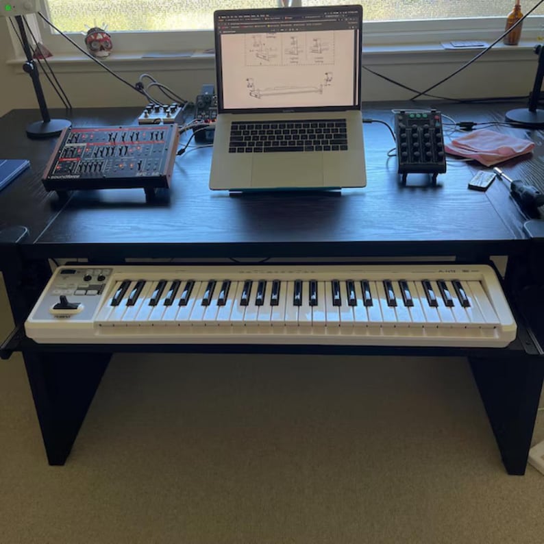 Puede incluir: Un escritorio negro con un teclado blanco, un ordenador port&aacute;til y equipos de audio. El teclado est&aacute; en primer plano, con un ordenador port&aacute;til que muestra un diagrama t&eacute;cnico en el centro. Otros dispositivos de audio est&aacute;n en el escritorio.
