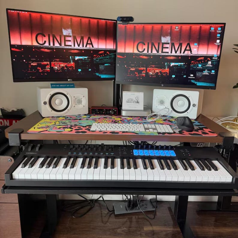 Puede incluir: Una configuraci&oacute;n de estudio en casa con dos monitores de computadora que muestran la palabra "CINEMA" en rojo. El escritorio incluye altavoces blancos, un teclado, un rat&oacute;n y un teclado MIDI. Una alfombrilla de rat&oacute;n colorida a&ntilde;ade un toque vibrante al espacio de trabajo.