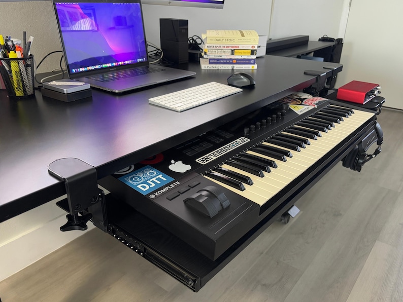 Puede incluir: Un escritorio negro con un port&aacute;til, un teclado y un teclado MIDI. El port&aacute;til est&aacute; abierto, mostrando una pantalla p&uacute;rpura. El teclado MIDI est&aacute; montado debajo del escritorio y tiene teclas negras y crema. El escritorio tambi&eacute;n tiene un rat&oacute;n y una pila de libros.