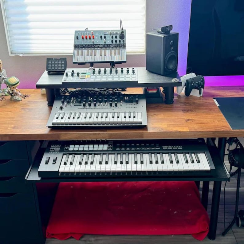 Puede incluir: Una configuraci&oacute;n de estudio en casa con varios sintetizadores y equipos de audio en un escritorio de madera. La configuraci&oacute;n incluye un altavoz negro, una peque&ntilde;a mesa de mezclas y un teclado. Se ve un coj&iacute;n rojo debajo del escritorio.
