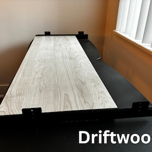 Puede incluir: Una mesa rectangular con una superficie de veta de madera clara y soportes de metal negro. La mesa est&aacute; etiquetada "Driftwood" en texto blanco en la parte inferior. La mesa est&aacute; colocada contra una pared neutra y cerca de una ventana con persianas blancas.