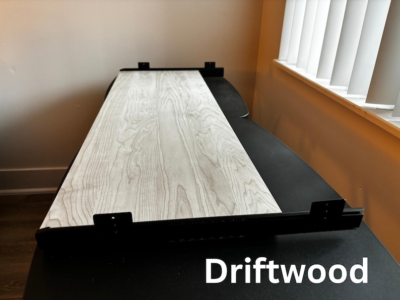 Puede incluir: Una mesa rectangular con una superficie de veta de madera clara y soportes de metal negro. La mesa est&aacute; etiquetada "Driftwood" en texto blanco en la parte inferior. La mesa est&aacute; colocada contra una pared neutra y cerca de una ventana con persianas blancas.