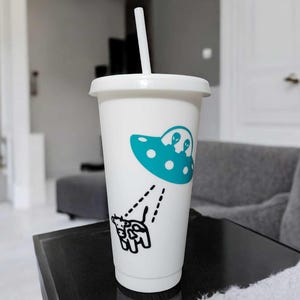 May include: White travel cup with a lid and straw. The cup features a teal UFO graphic with two alien figures and a black and white cow being abducted by the UFO. The cup is on a black surface.