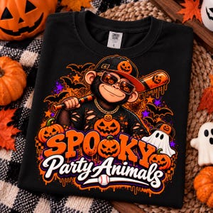 May include: A black t-shirt featuring a graphic of a monkey wearing a baseball cap and sunglasses, holding a baseball bat. The design includes pumpkins, a ghost, bats, and the text "SPOOKY Party Animals" in orange and white lettering.