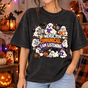 Puede incluir: Una camiseta gris carbón con un gráfico de varios fantasmas de dibujos animados realizando diversas actividades de comunicación. El texto dice: "HOWEVER YOU COMMUNICATE, I AM LISTENING." El diseño incluye calabazas naranjas y murciélagos.