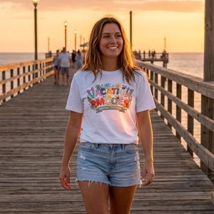 May include: A woman walks on a wooden pier towards the viewer, wearing a white t-shirt with the words "Vacation Mode" in colorful text, denim shorts, and sandals. The background shows the ocean and a sunset.