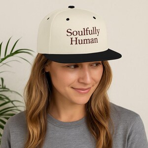 Peut inclure: Casquette de baseball à visière plate crème avec une visière et un bouton supérieur noirs. Le texte "Soulfully Human" est imprimé en bordeaux sur le devant de la casquette. La casquette est portée par une personne.
