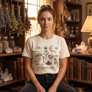 Peut inclure: Un t-shirt crème avec un motif graphique fantaisiste comprenant une fleur oculaire, un champignon, une clé, un scarabée et un fantôme dans une tasse à thé. Le texte "Tiny Oddities Cabinet" est imprimé en dessous. La femme est assise devant une bibliothèque.