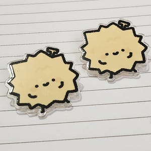 May include: Two cartoon-style durian fruit earrings. Each earring features a light yellow durian with a black outline and a smiling face. The earrings have a clear, shiny border and are set against a lined paper background.
