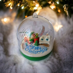 May include: A clear glass snow globe ornament with a green base. Inside the globe is a miniature Christmas scene with a red truck, a house, and a sign that reads "Spirit of St. Nick". The base of the globe has the words "Merry Christmas" written on it.